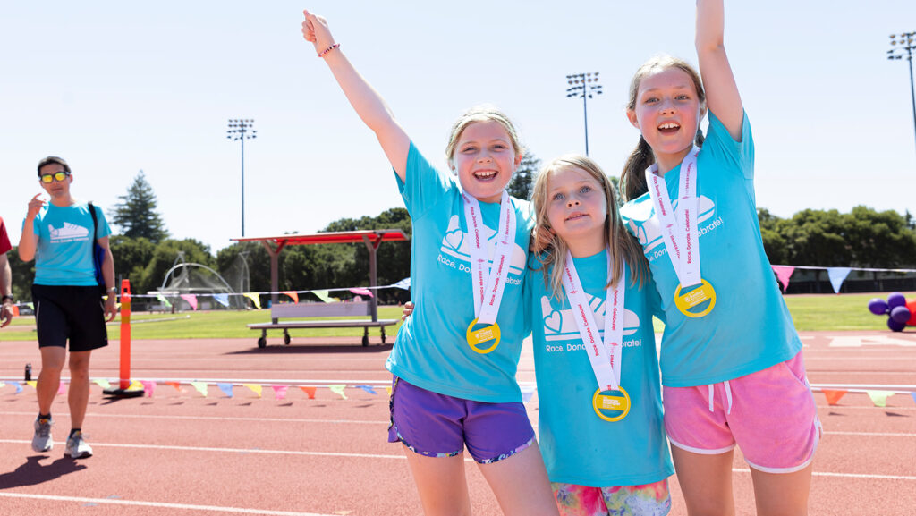 Three girls with summer scamper shirts on and race medals pose on the Stanford track.