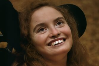 A smiling young woman with long brown hair looks upward, her face warmly lit against a softly blurred, earthy-toned background.