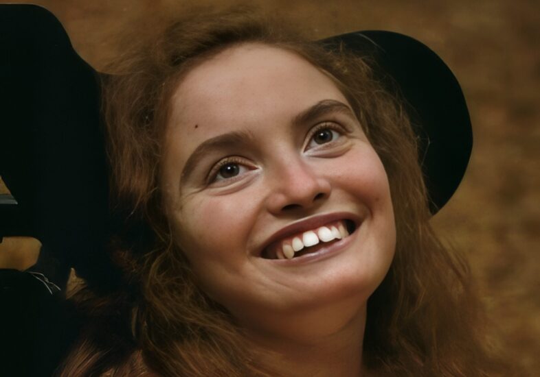 A smiling young woman with long brown hair looks upward, her face warmly lit against a softly blurred, earthy-toned background.