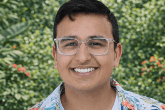 Smiling young man wearing clear-framed glasses and a white floral button-up shirt over a light T-shirt, standing outdoors in front of a lush green garden background.