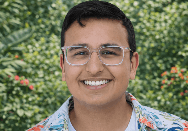 Smiling young man wearing clear-framed glasses and a white floral button-up shirt over a light T-shirt, standing outdoors in front of a lush green garden background.