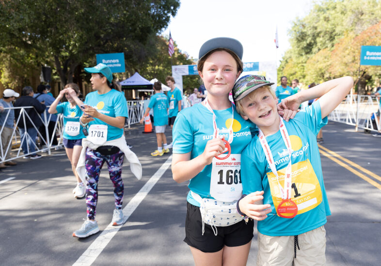 Two young children in teal Summer Scamper shirts grin at the camera after finishing the race, displaying their medals. The older child has an arm around the younger one, who holds up a Kids Fun Run bib.