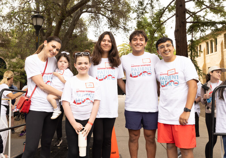 A group of six people — a woman holding a child, two children, and two young adults — pose together wearing white "Summer Scamper Patient Hero" t-shirts at the event.